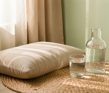 Coin de méditation lumineux avec coussin neutre, verre d’eau à température ambiante et bouteille en verre dans une ambiance douce et naturelle.