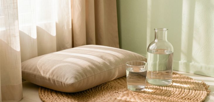 Coin de méditation lumineux avec coussin neutre, verre d’eau à température ambiante et bouteille en verre dans une ambiance douce et naturelle.